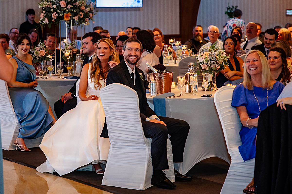 bride and groom laughing during toasts at their wedding reception, surrounded by their guests