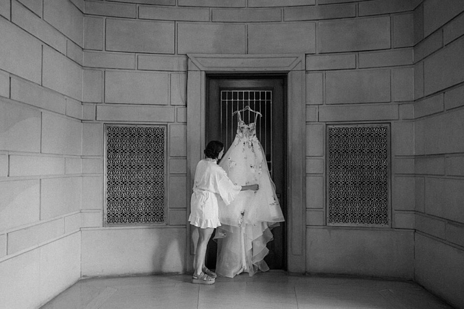 A bride in a white robe stands in front of a door, touching a wedding dress that is hanging on the door of the lobby at Stambaugh Auditorium.